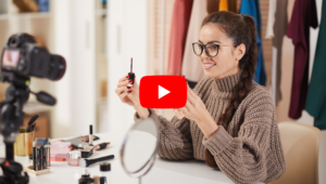 A young woman wearing glasses holds mascara in her hands and is showing it to a camera on the left. The YouTube logo is in the center of the image. With the video QR code, you can easily share any video.